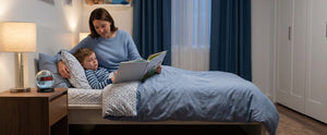 A woman sits in bed with a child reading a book in a cozy bedroom with blue curtains, a bedside alarm clock, and a wooden nightstand.