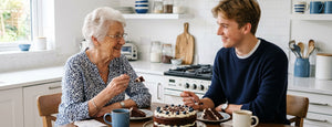 An elderly woman and a young man sitting at a kitchen table sharing a moment, with a chocolate cake in the foreground and a modern white kitchen in the background.
