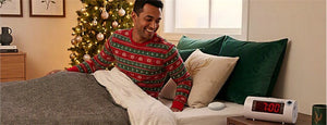 A man wearing a festive red and green Christmas sweater sits up in bed next to a glowing digital clock, with a decorated Christmas tree and green pillows in the background.