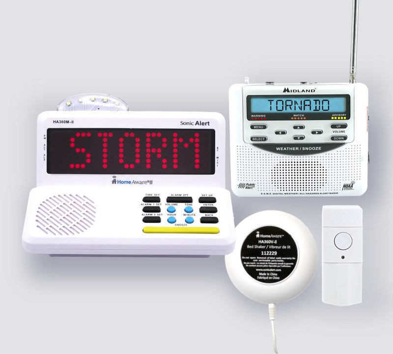 A storm warning kit including a white HomeAware II main unit with a red LED 'STORM' display, a white radio receiver with a blue LCD screen showing 'TORNADO', a white round bed shaker, and a security alert transmitter arranged on a light gray background.
